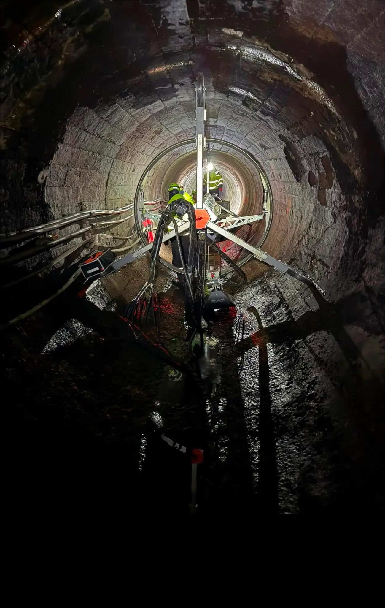 Arbejdshold med udstyr dybt inde i en tunnel, oplyst af arbejdslys mod de våde murstensvægge.
